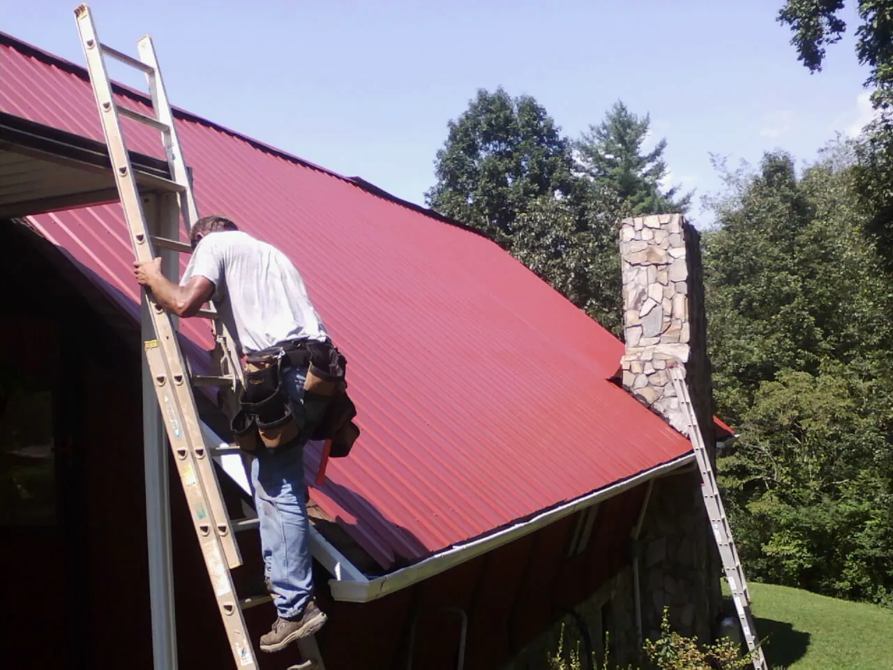 Roofer on a ladder beside a red metal roof and stone chimney — Soffit & Fascia Repair by Roofing Brothers in Winder